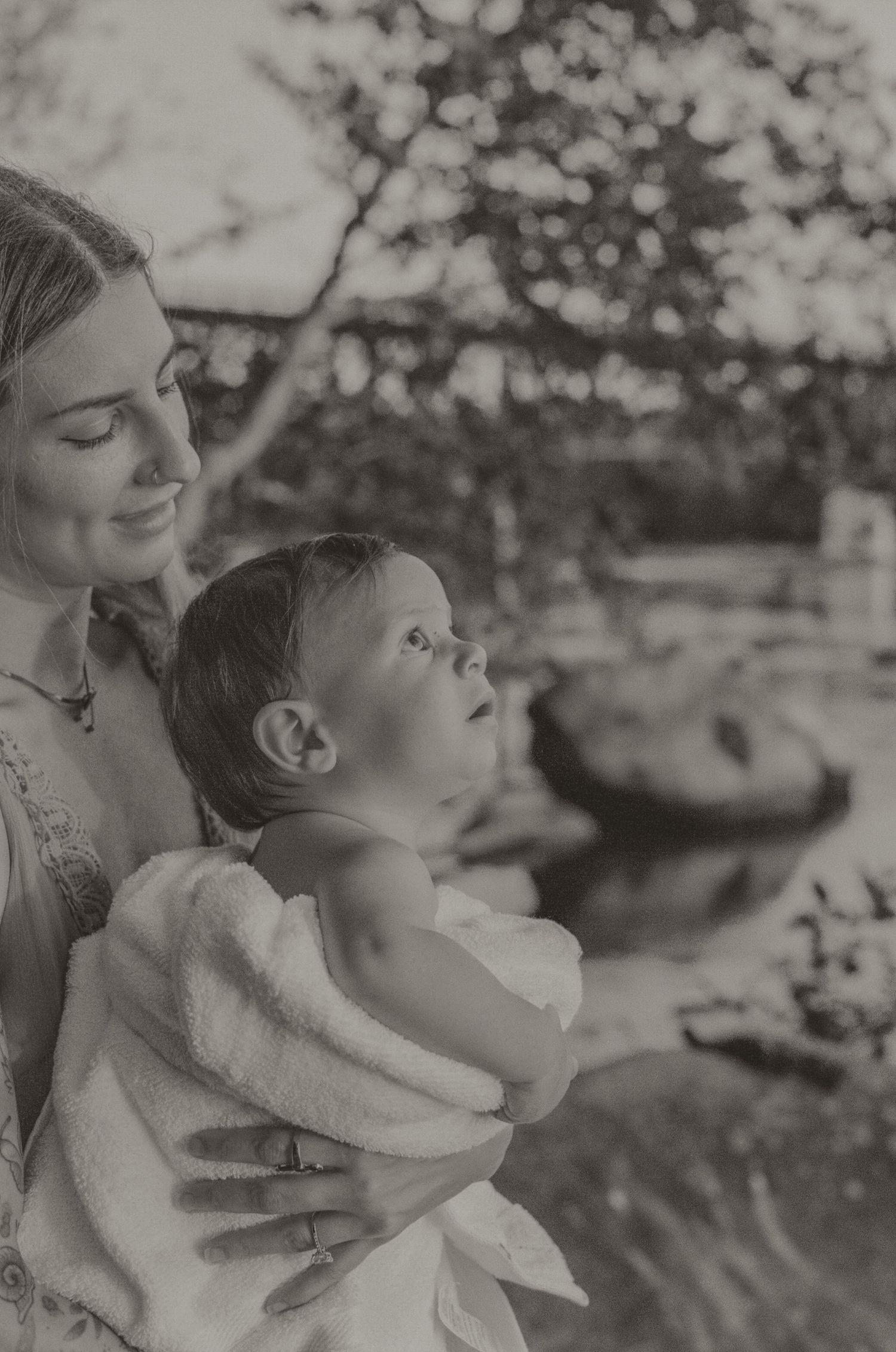 Black and white photo of a smiling mother holding her baby wrapped in a white towel outdoors.
