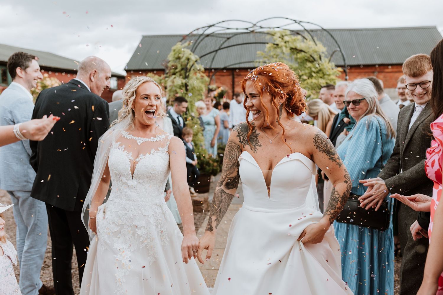 Two brides in white wedding gowns walk through confetti shower, celebrating their same-sex wedding ceremony outdoors.