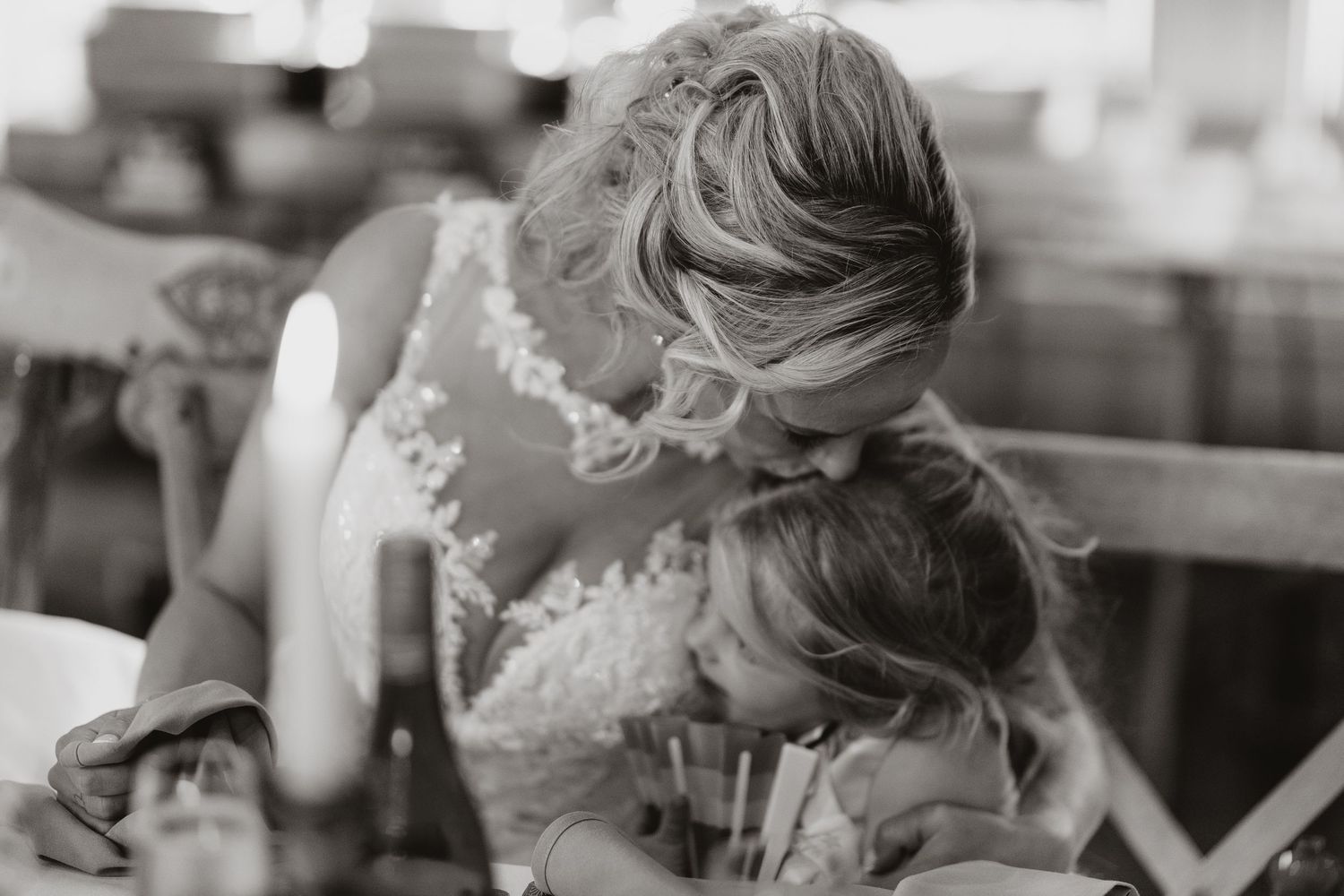 Bride in lace gown leaning down to kiss a young flower girl at a wedding reception.