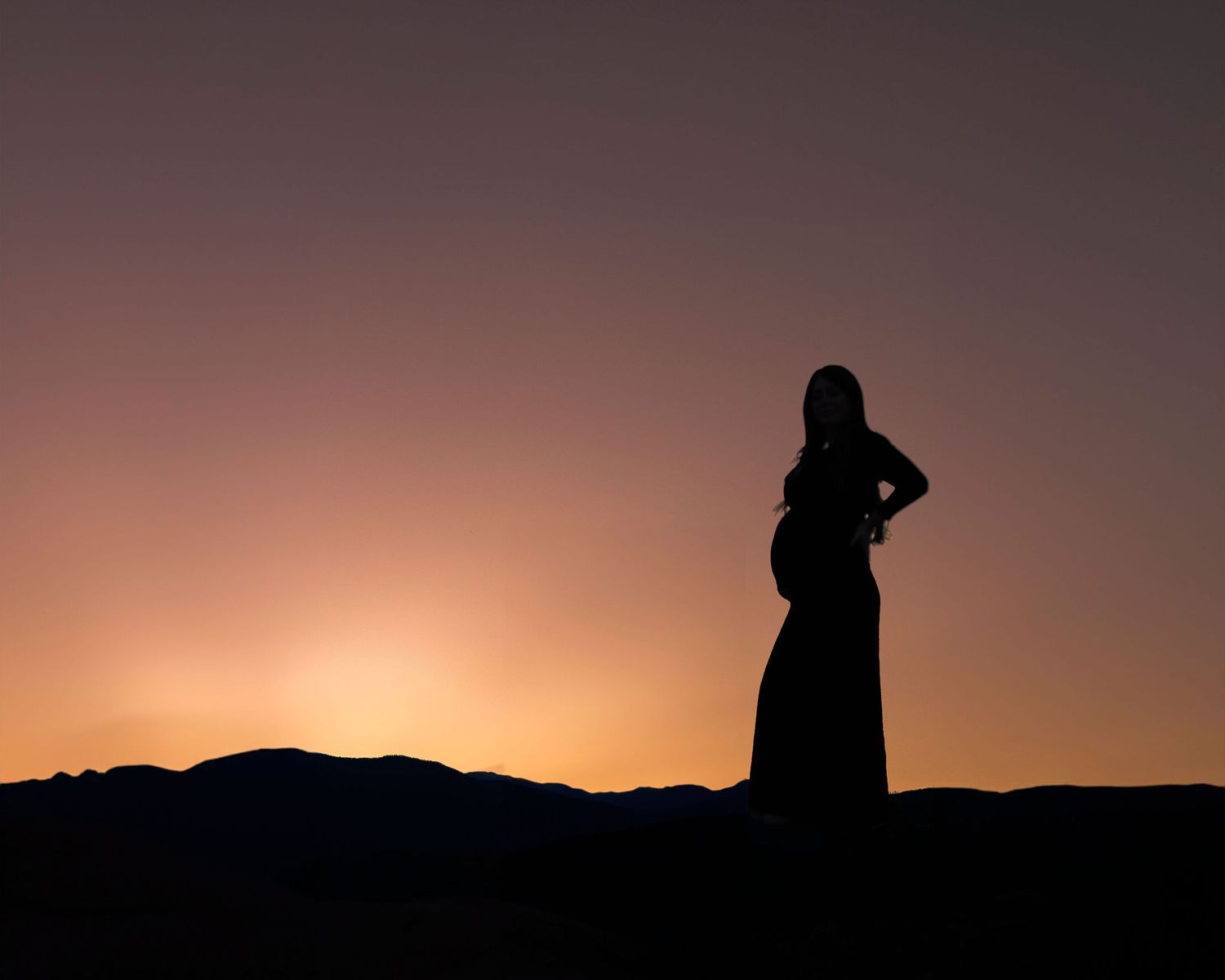 Silhouette of a pregnant woman standing against a vibrant sunset sky with mountain horizon.