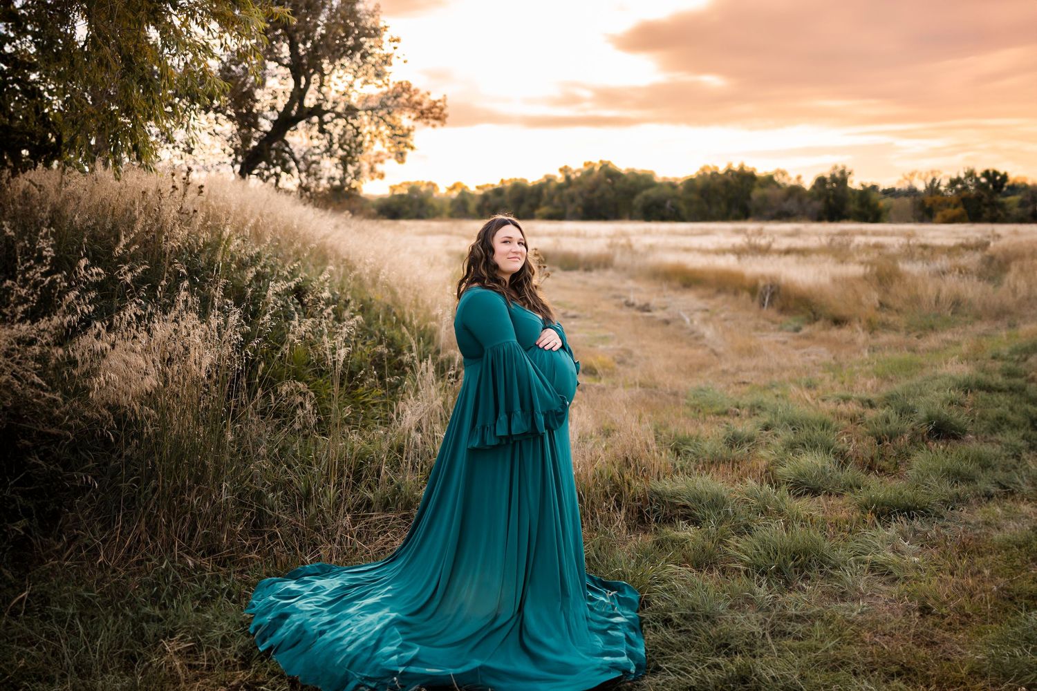 Pregnant woman in teal gown standing in golden field at sunset.