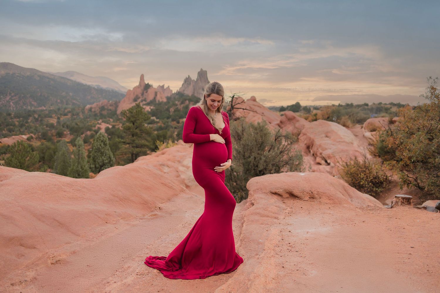 Pregnant woman in red maternity gown poses on red rocks at Garden of the Gods at sunset.