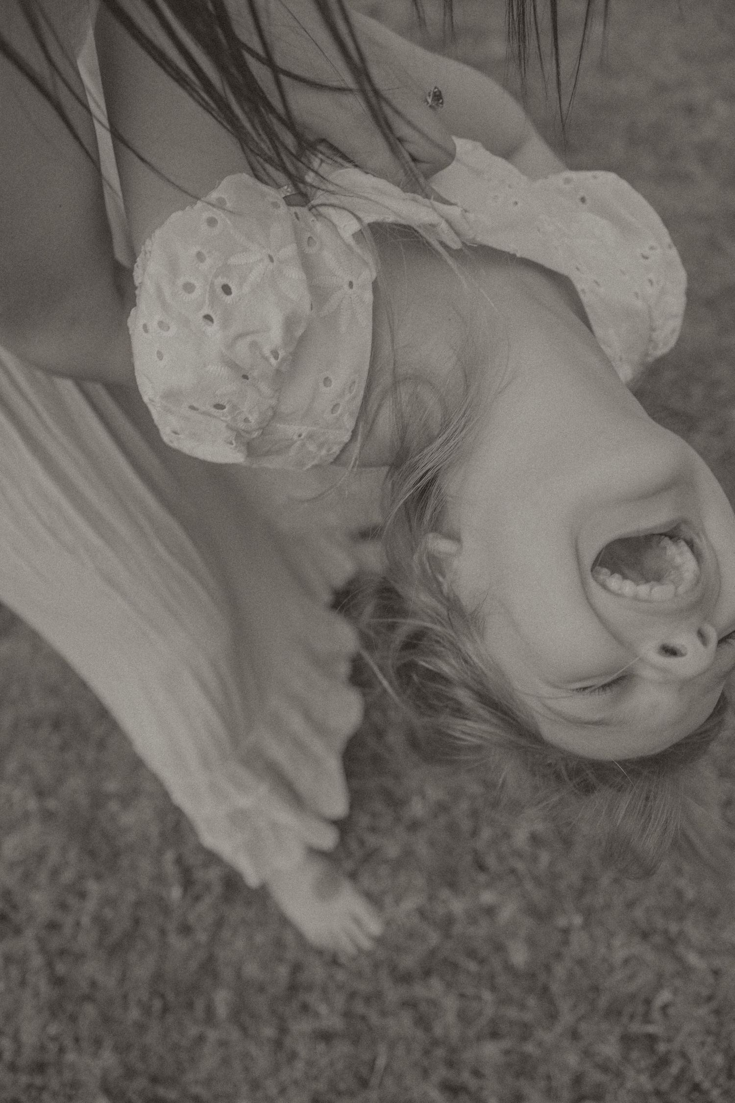 Black and white photo of a smiling child hanging upside down outdoors, wearing a white eyelet dress.