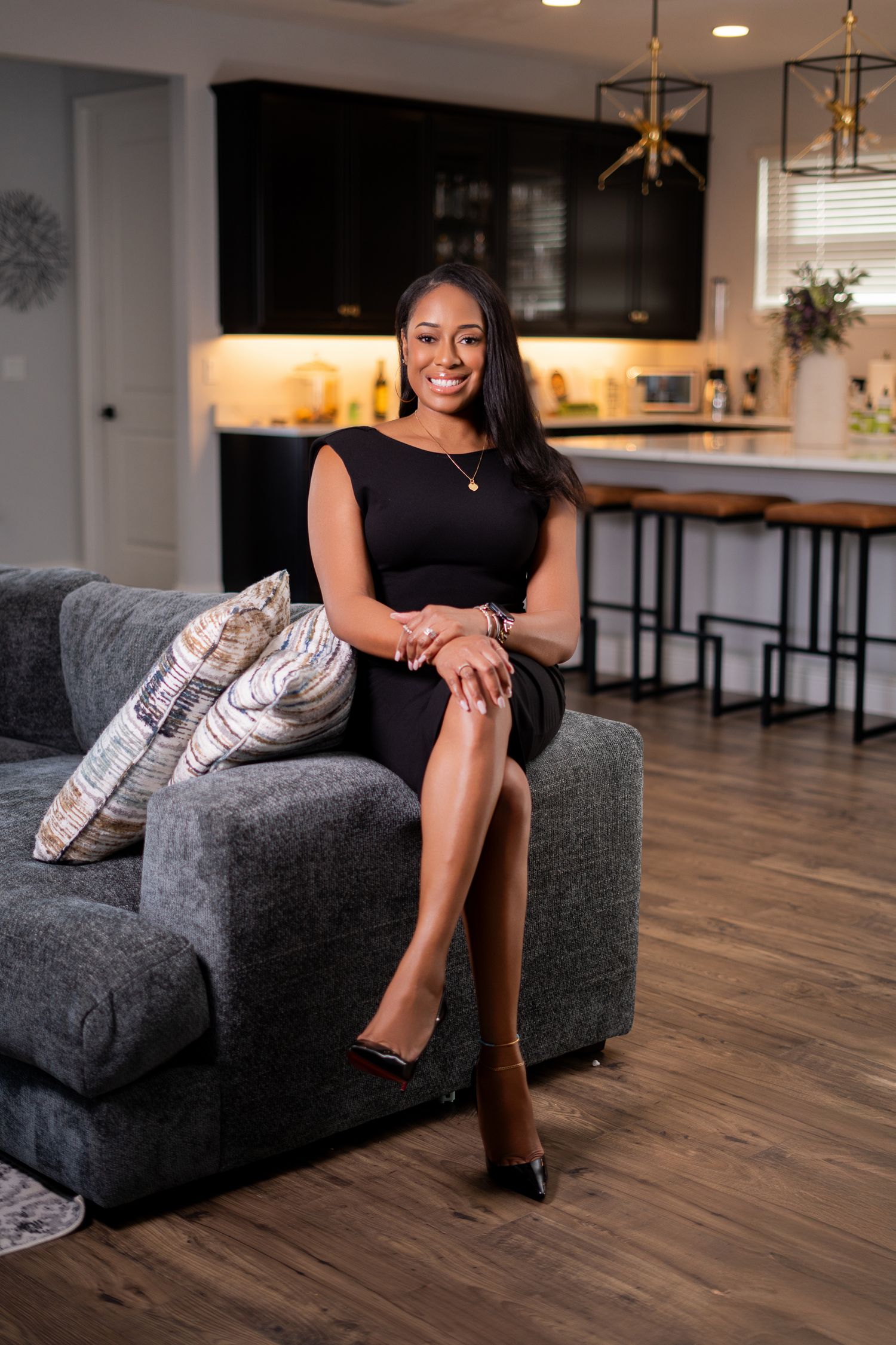 A smiling woman in a black dress sits elegantly on a gray sofa in a modern open-concept kitchen and living area.