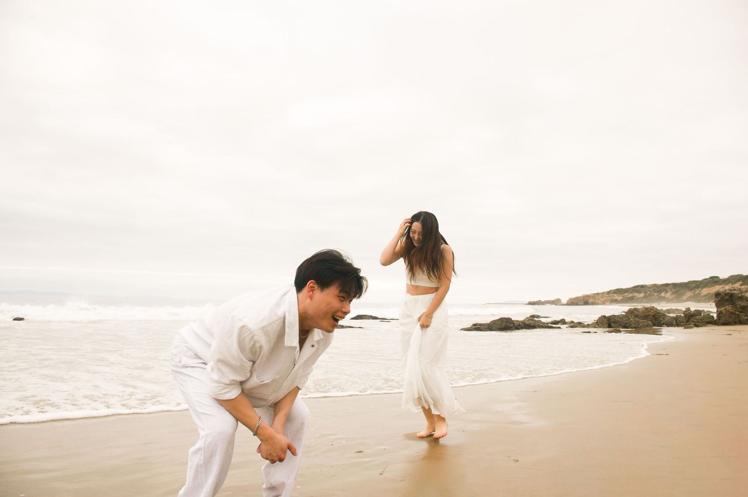 Couple in white outfits playing on a sandy beach with ocean waves behind them.