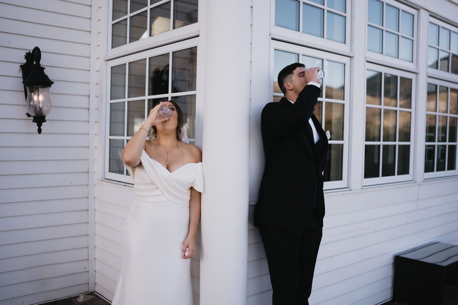Bride in white off-shoulder gown and groom in black tuxedo leaning against white building columns outdoors.