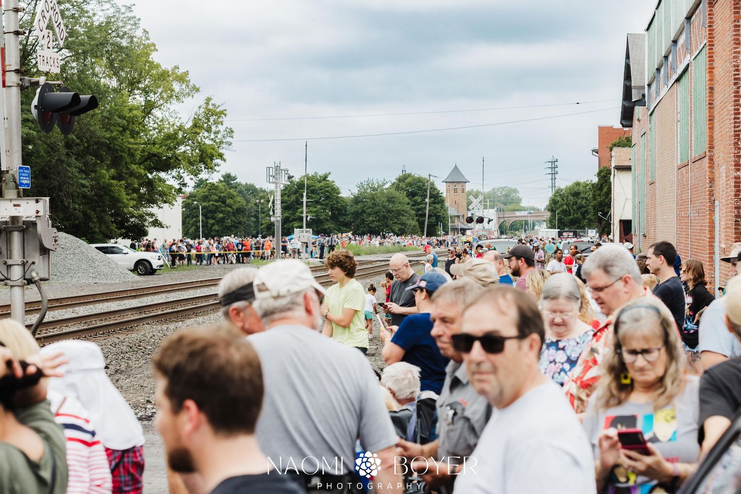Large crowd gathers near railroad tracks at an outdoor community event on a cloudy day in a small town.