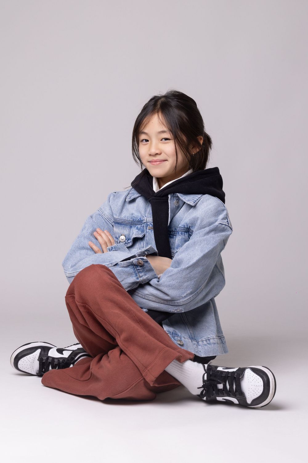 A model in denim jacket and brown pants poses casually on the floor in a studio setting against a white background.