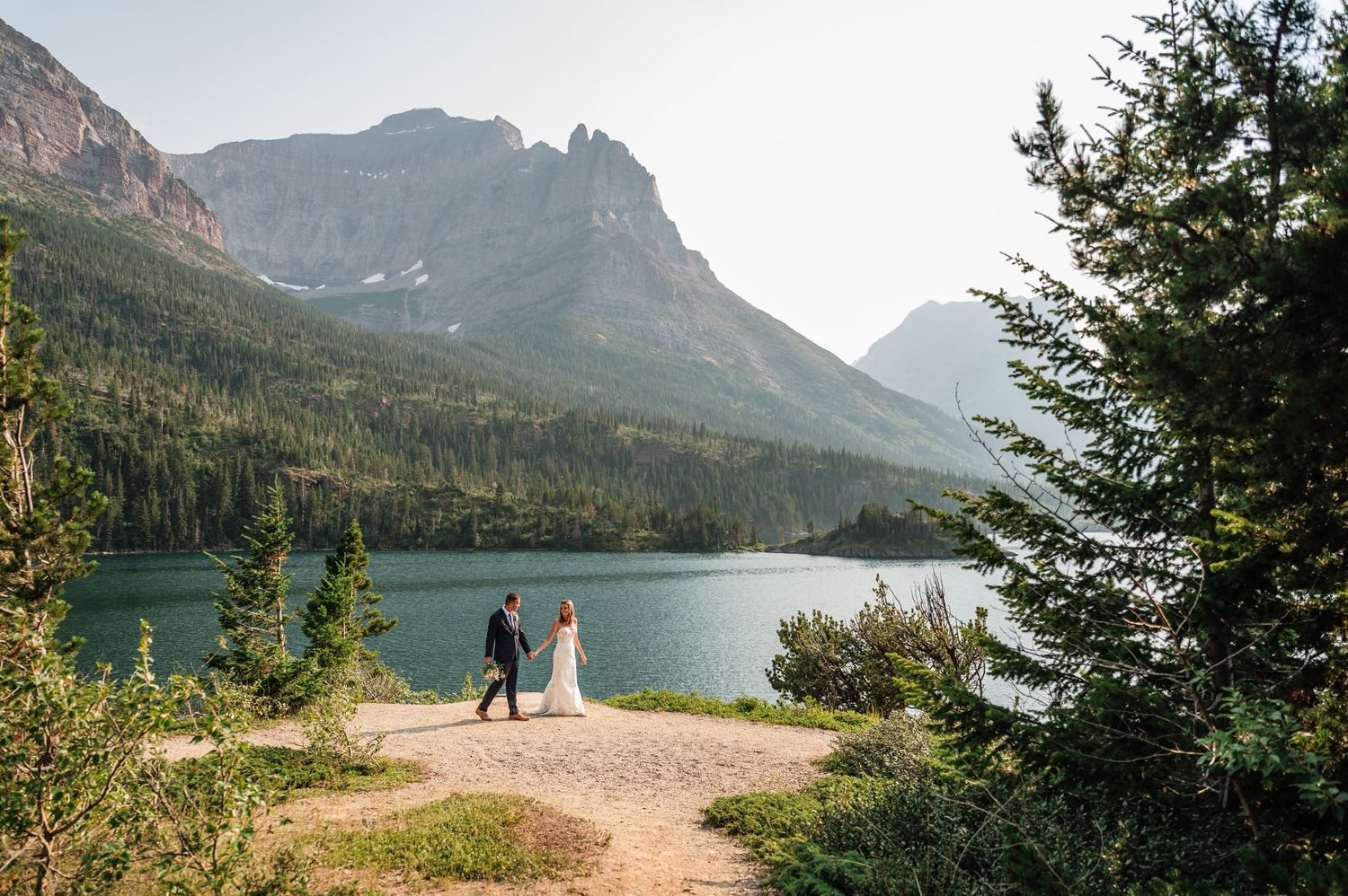 Couple holds hands on a lakeside trail with stunning mountain backdrop at Glacier National Park wedding.