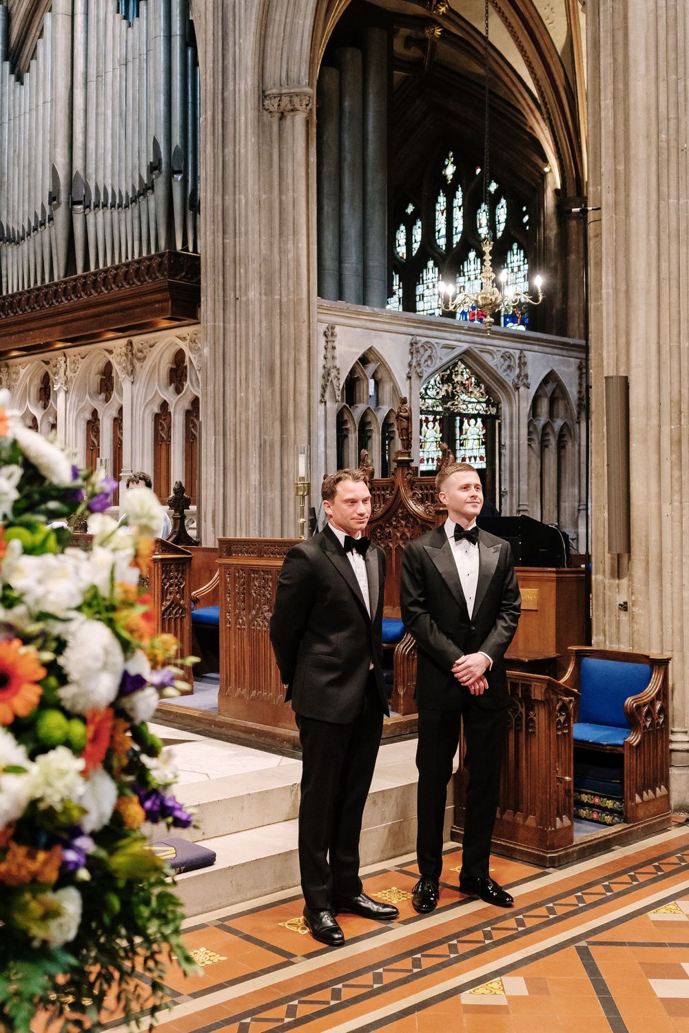 Groom in black tuxedos standing at St Mary Redcliffe Church altar with pipe organ, stained glass windows, and floral arrangements nearby.