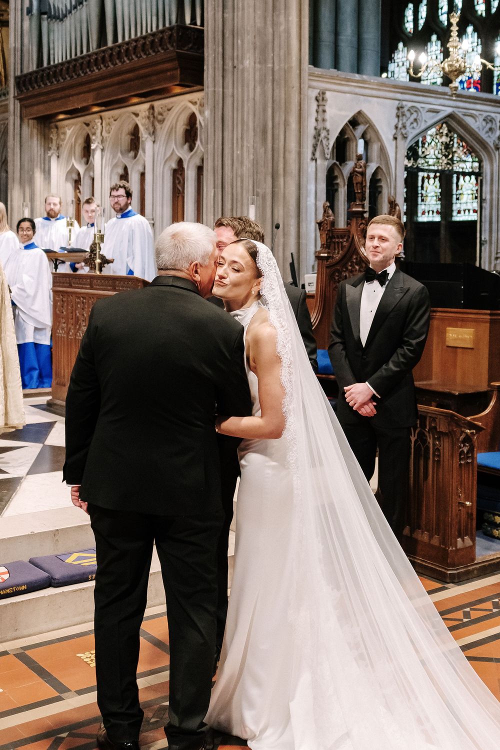 Bride in cathedral receives kiss from father as groom in tuxedo at St Mary Redcliffe Church, Bristol Wedding