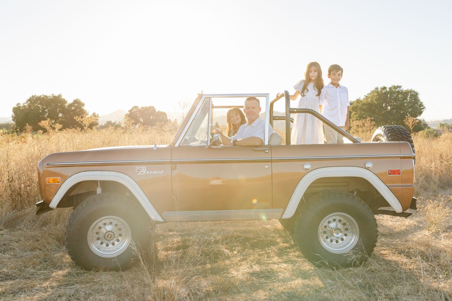 Family enjoying a sunny off-road adventure in a vintage Ford Bronco through golden dry grasslands.