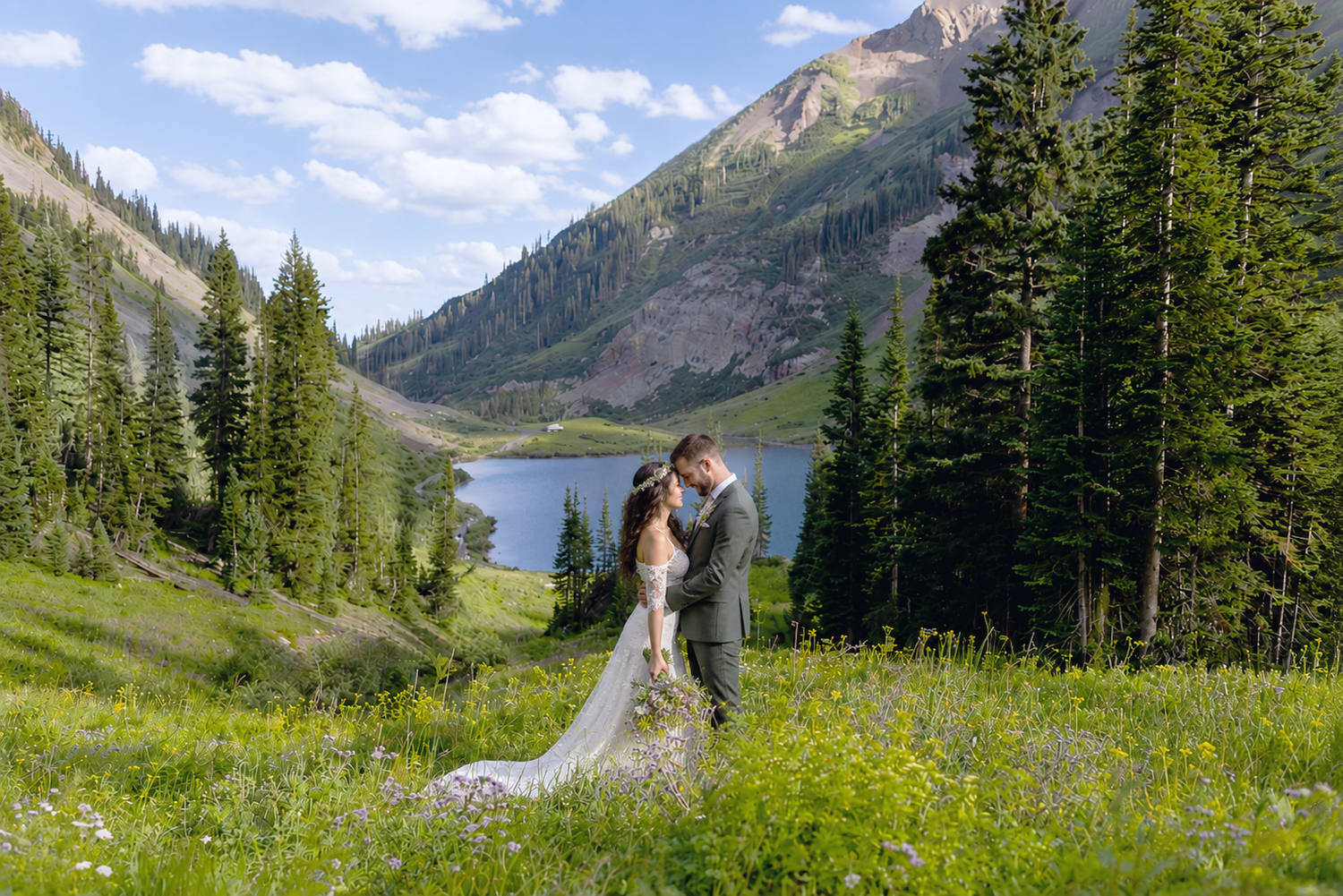 Bride and groom kissing in a wildflower meadow with a mountain lake and pine trees in the background.