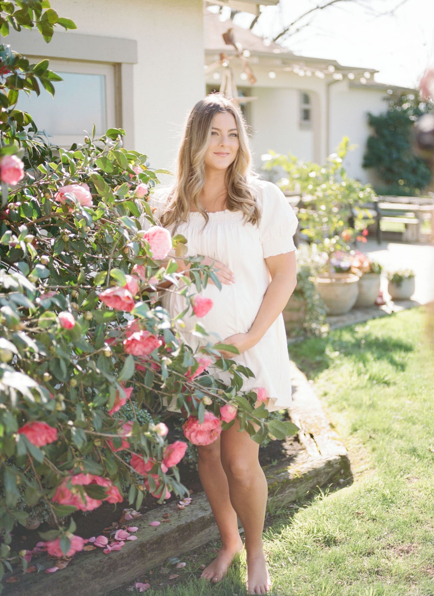 A woman in a white dress stands barefoot beside blooming pink roses in a sunny garden near a house.