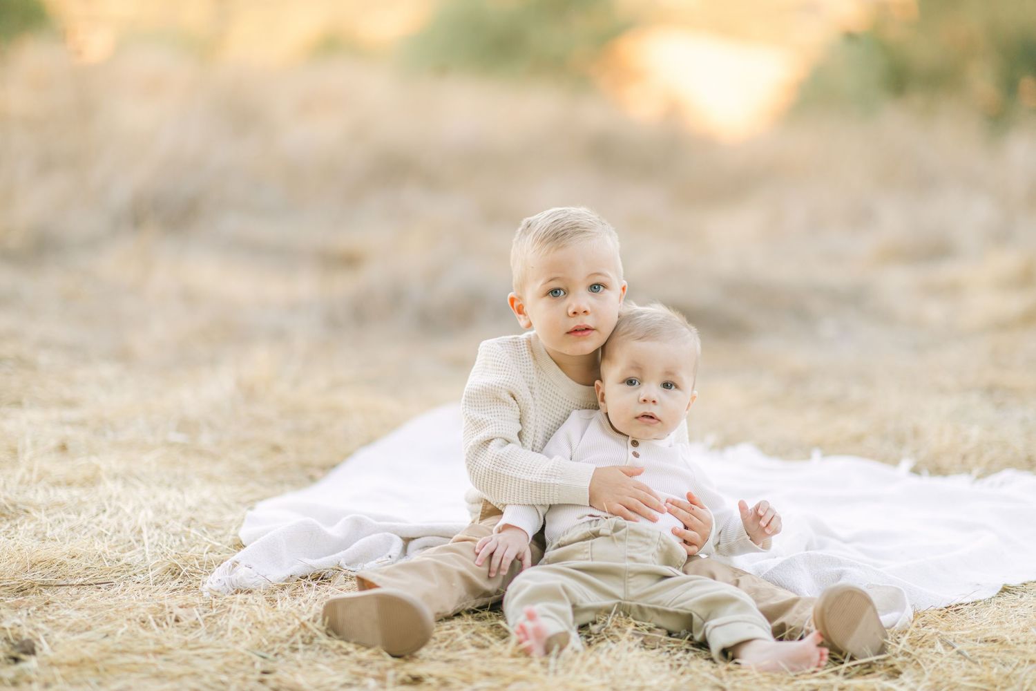Two young siblings in neutral outfits sit together on a white blanket in a golden field.