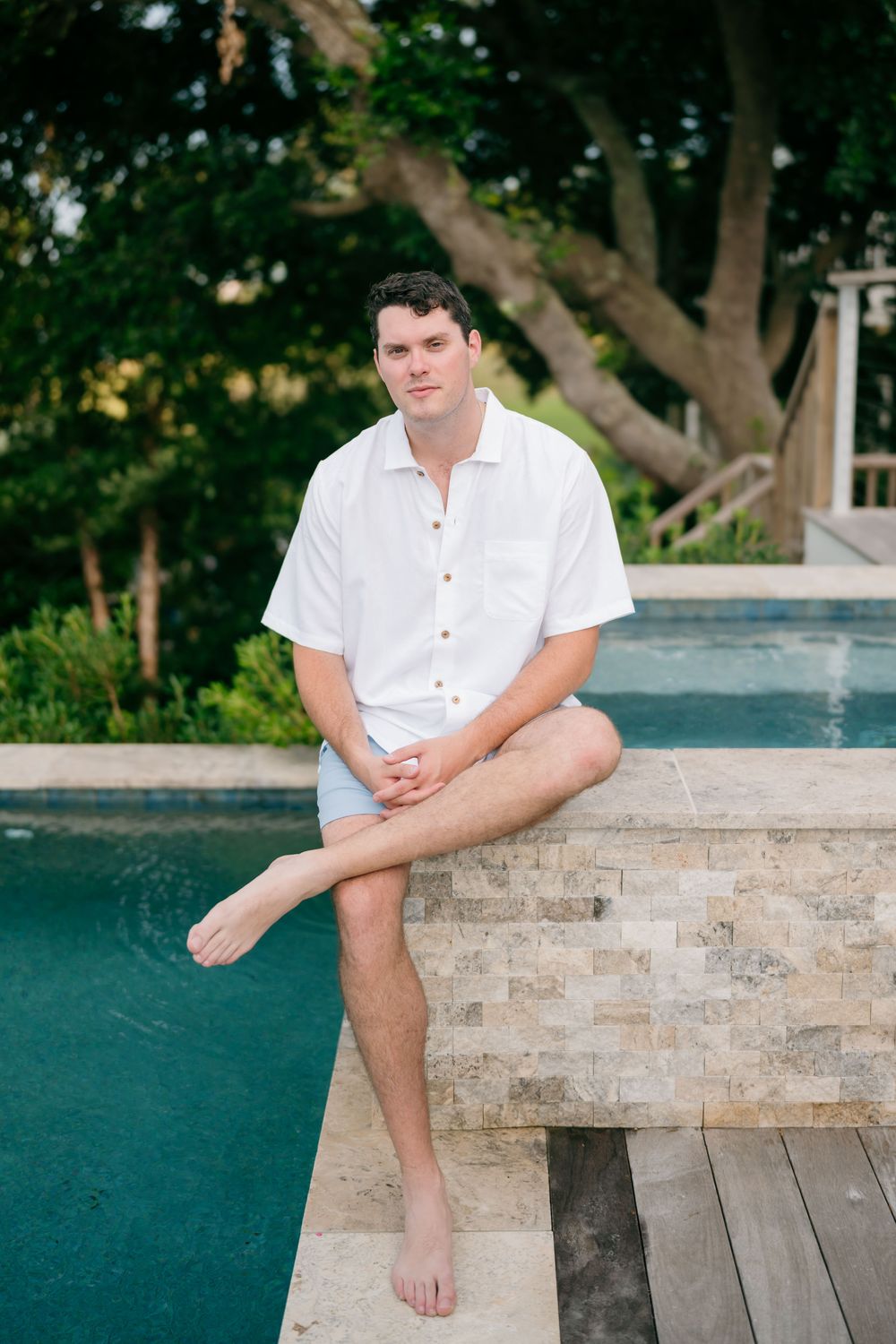 A young man in a white shirt and shorts sits barefoot on a stone pool edge surrounded by lush green trees.