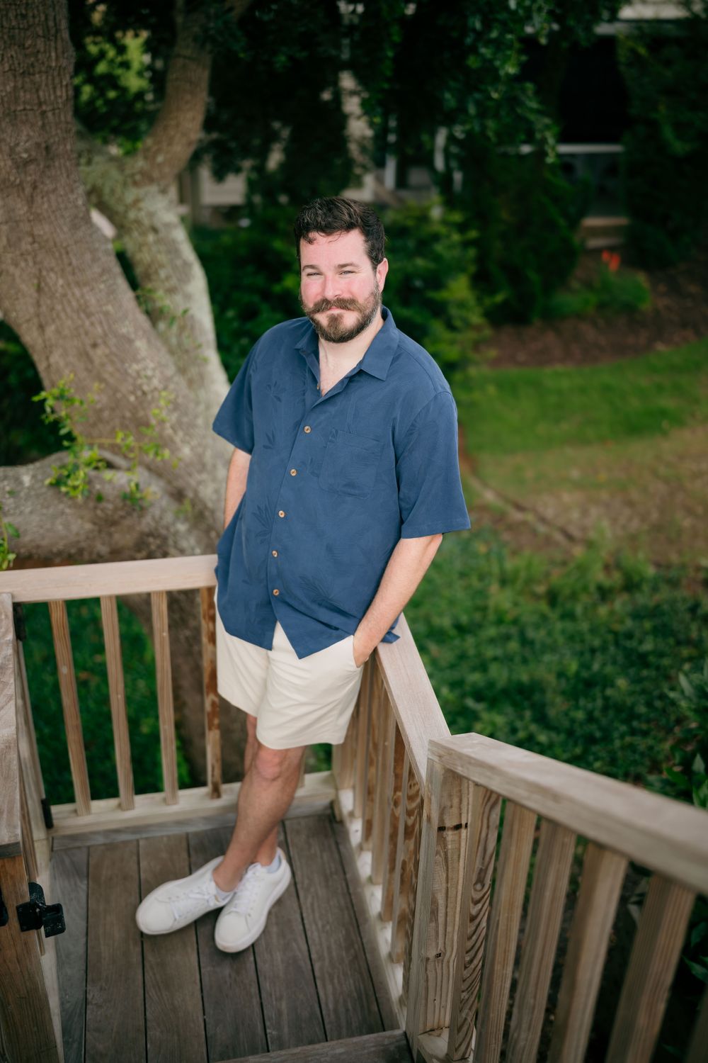 Smiling bearded man in navy blue shirt and cream shorts standing on a wooden deck surrounded by lush greenery.