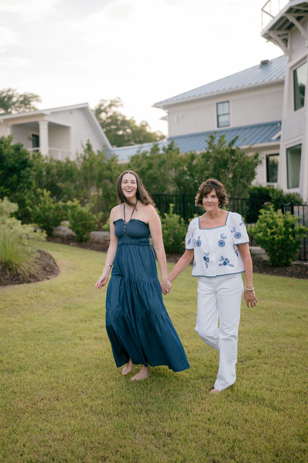 Two women holding hands walking on green lawn near white coastal home, one in teal maxi dress.