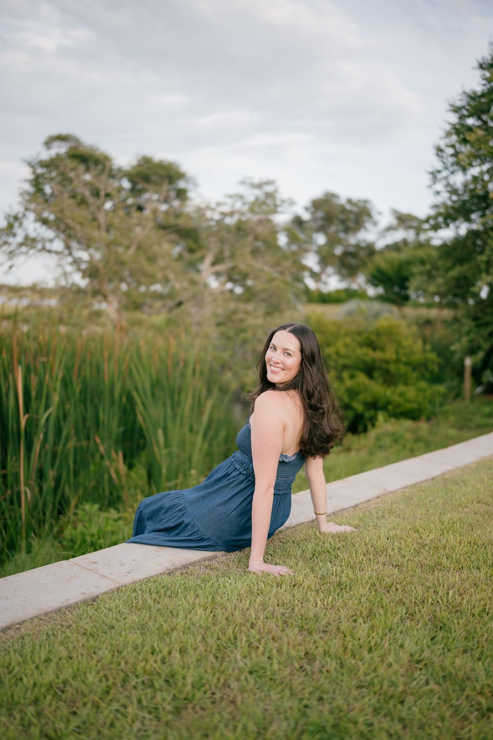 Young woman in blue dress sitting on grass edge smiling over shoulder with marsh and trees behind her.