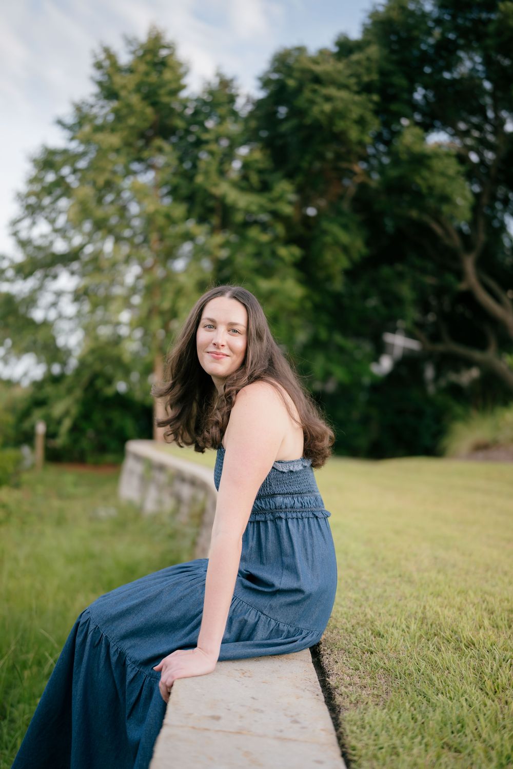 Smiling brunette woman in blue jumpsuit sitting on stone wall with lush green trees in background.