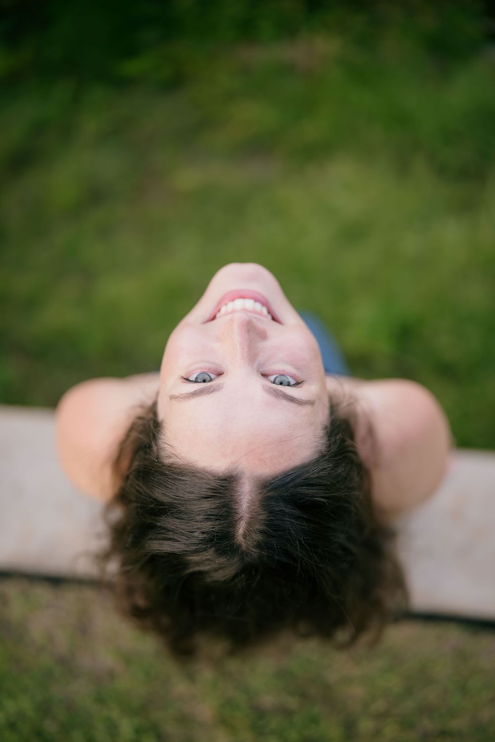Smiling woman with dark curly hair lying on grass, viewed from above, looking up with bright blue eyes.