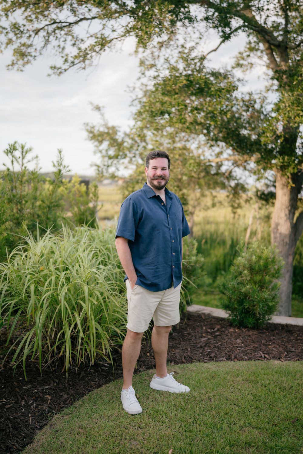 Man in navy shirt and khaki shorts smiling outdoors in a lush green garden setting.