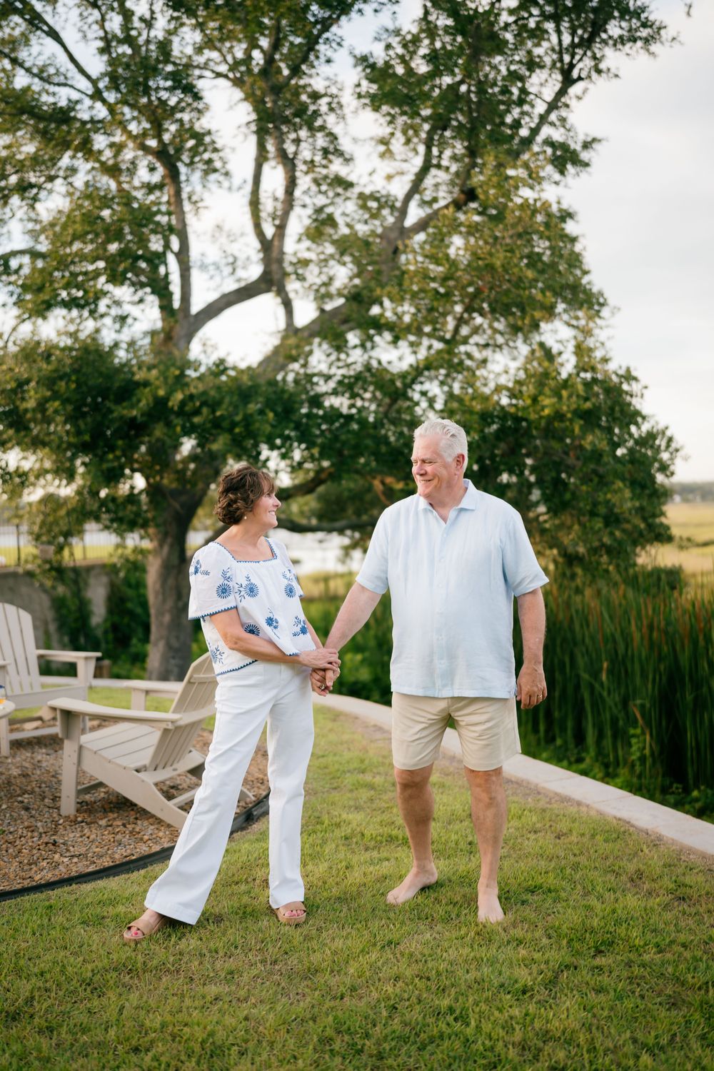 Older couple holding hands and walking barefoot on green lawn with marsh and trees in background.