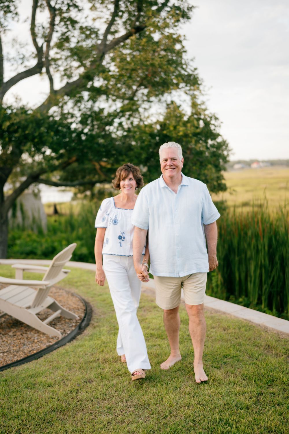 Happy older couple holding hands walking barefoot on green lawn with marsh views.