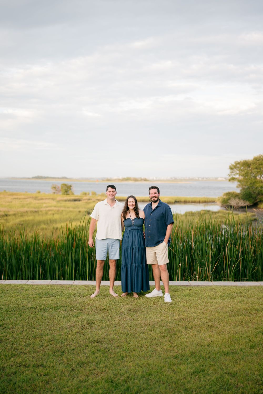 Three adults smiling together on green lawn with coastal marsh and water visible behind them.
