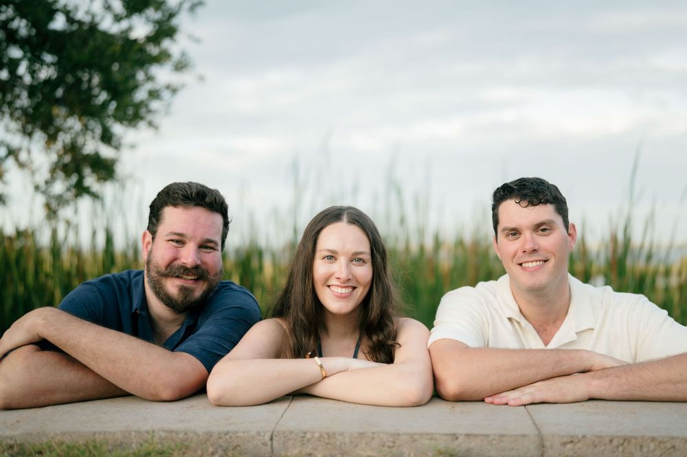Three smiling young adults posing outdoors leaning on a ledge with green grass and sky in the background.