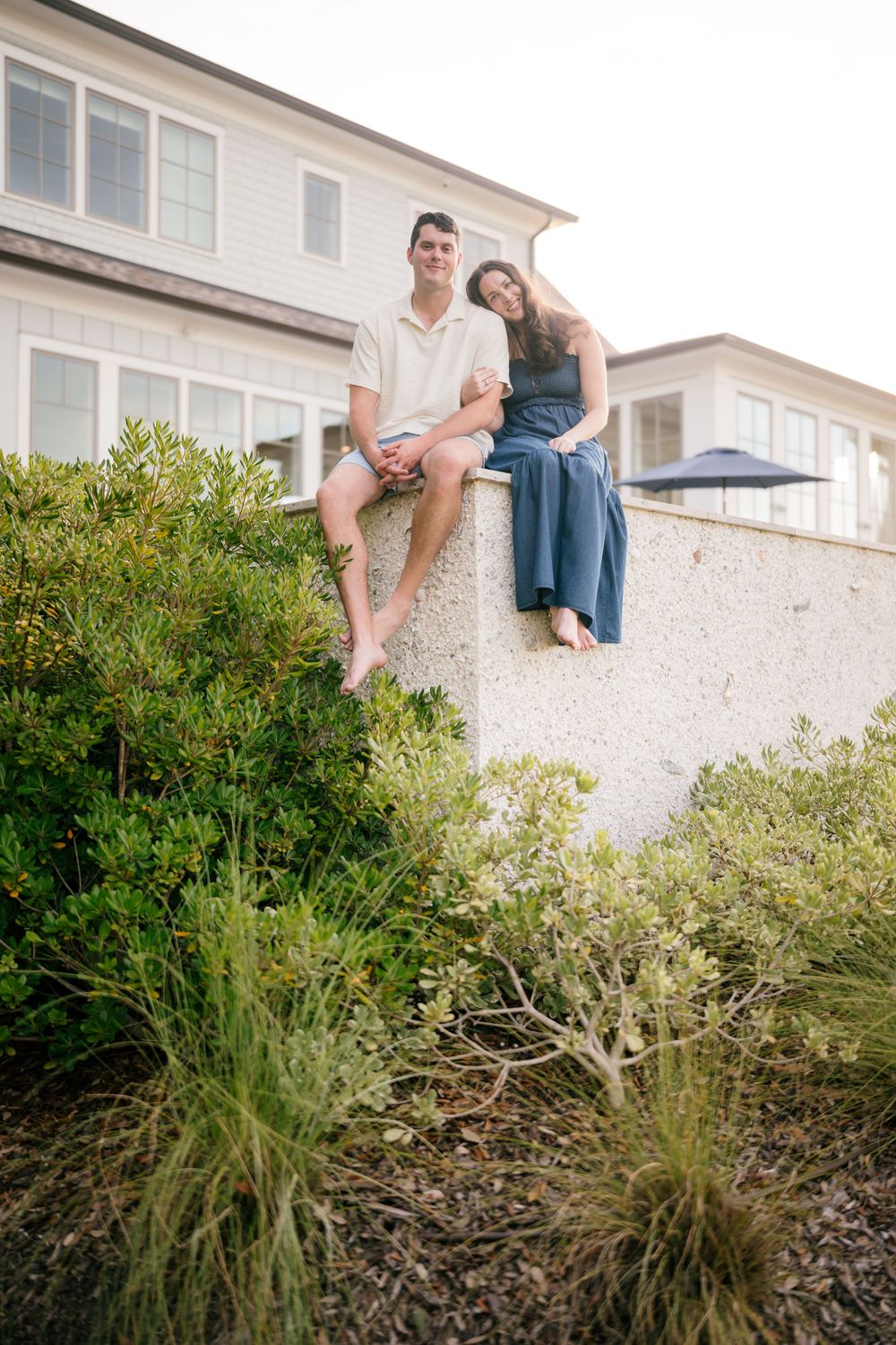 Young couple sitting barefoot on white stone wall surrounded by shrubs with beach house behind them.