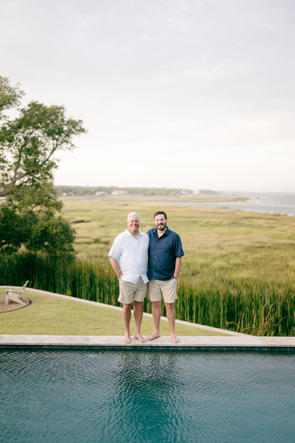 Two men standing barefoot by infinity pool with scenic marsh and waterway views in background.