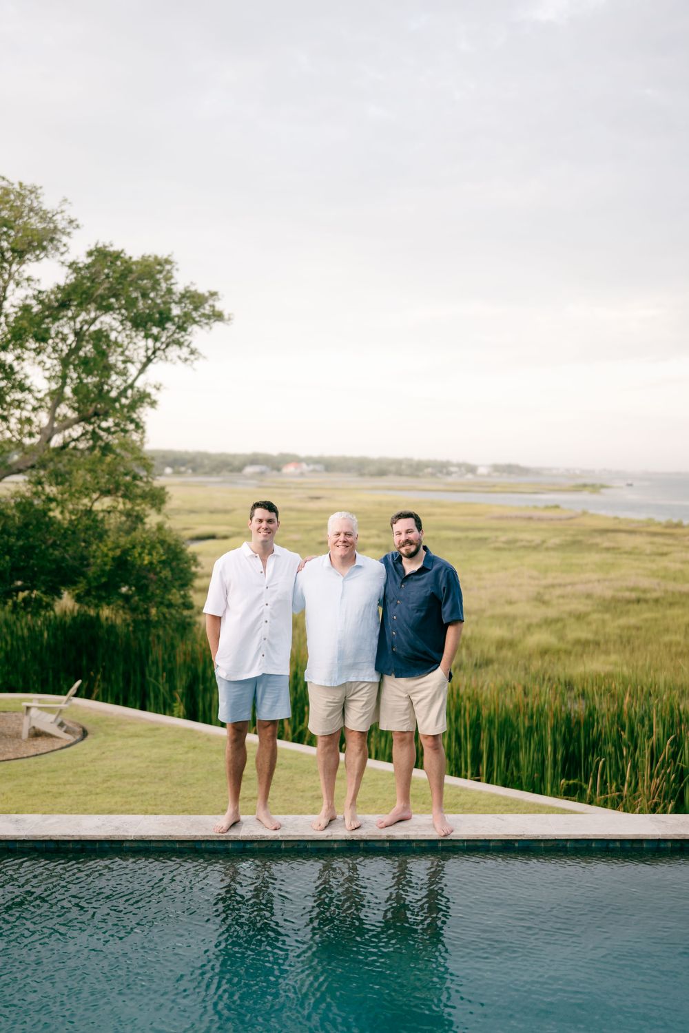 Three men smiling together barefoot by an infinity pool with expansive green marsh and coastal water views.