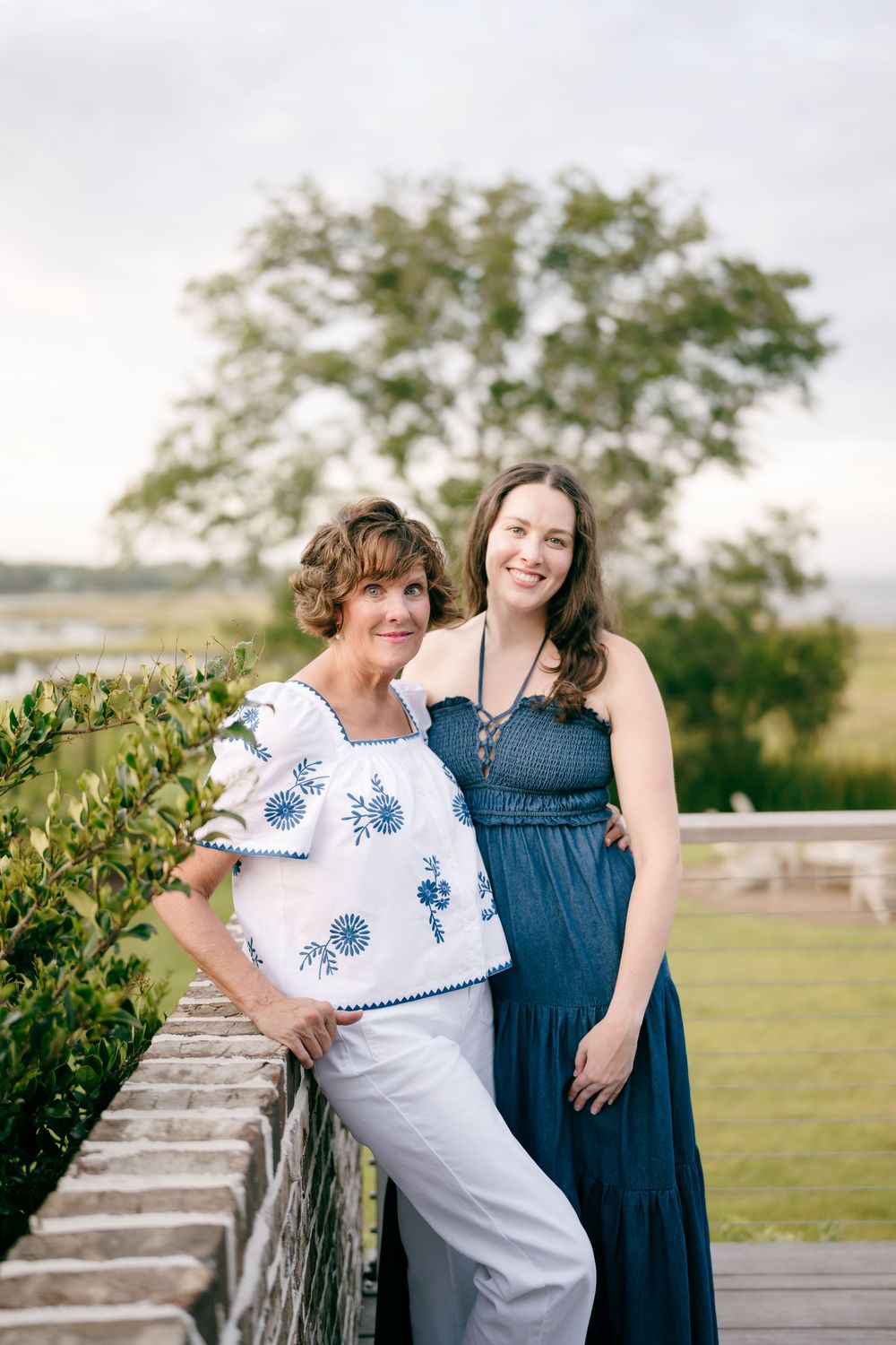 Two women smiling together on a wooden deck overlooking a marsh landscape on a cloudy day.