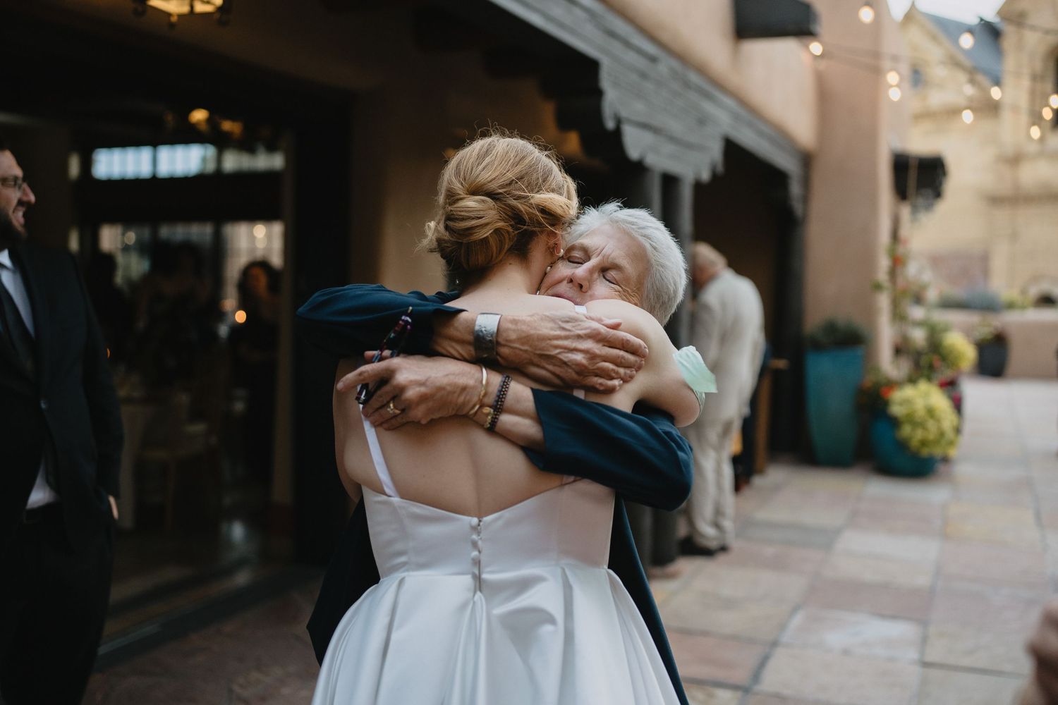 A bride in a white gown shares a heartfelt hug with an elderly woman at an outdoor wedding reception.