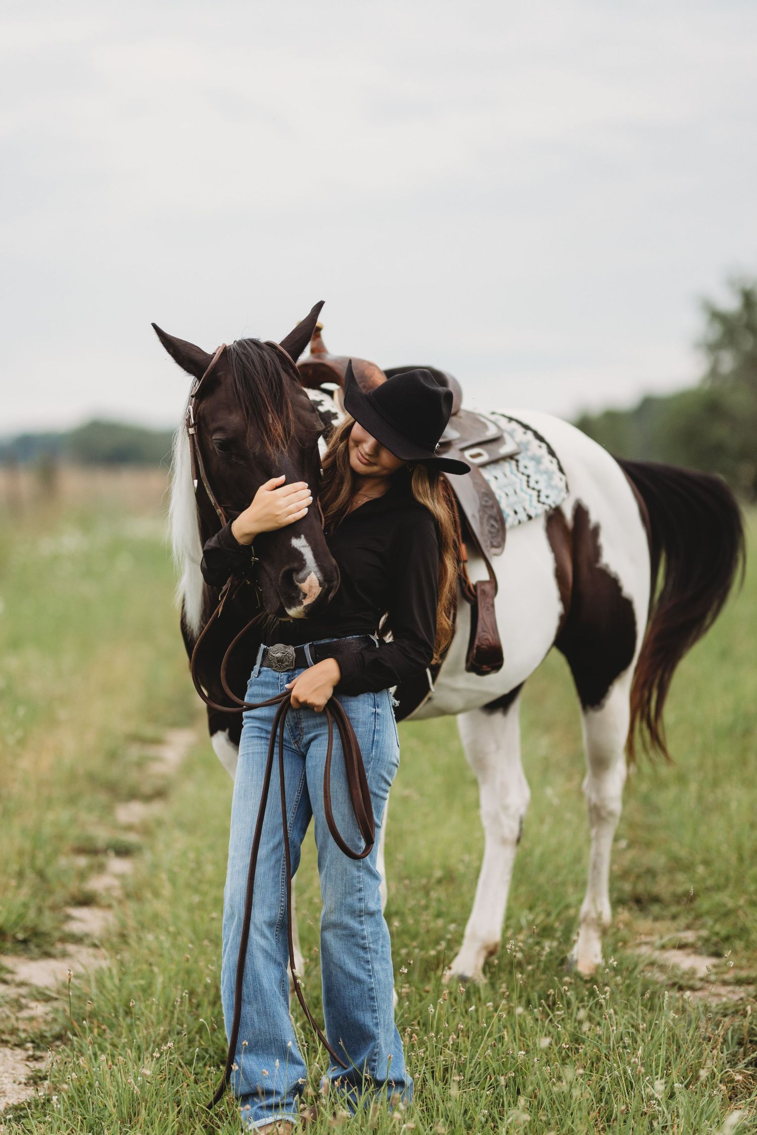 A woman in a black hat and jeans affectionately embraces a brown and white paint horse in a green field.