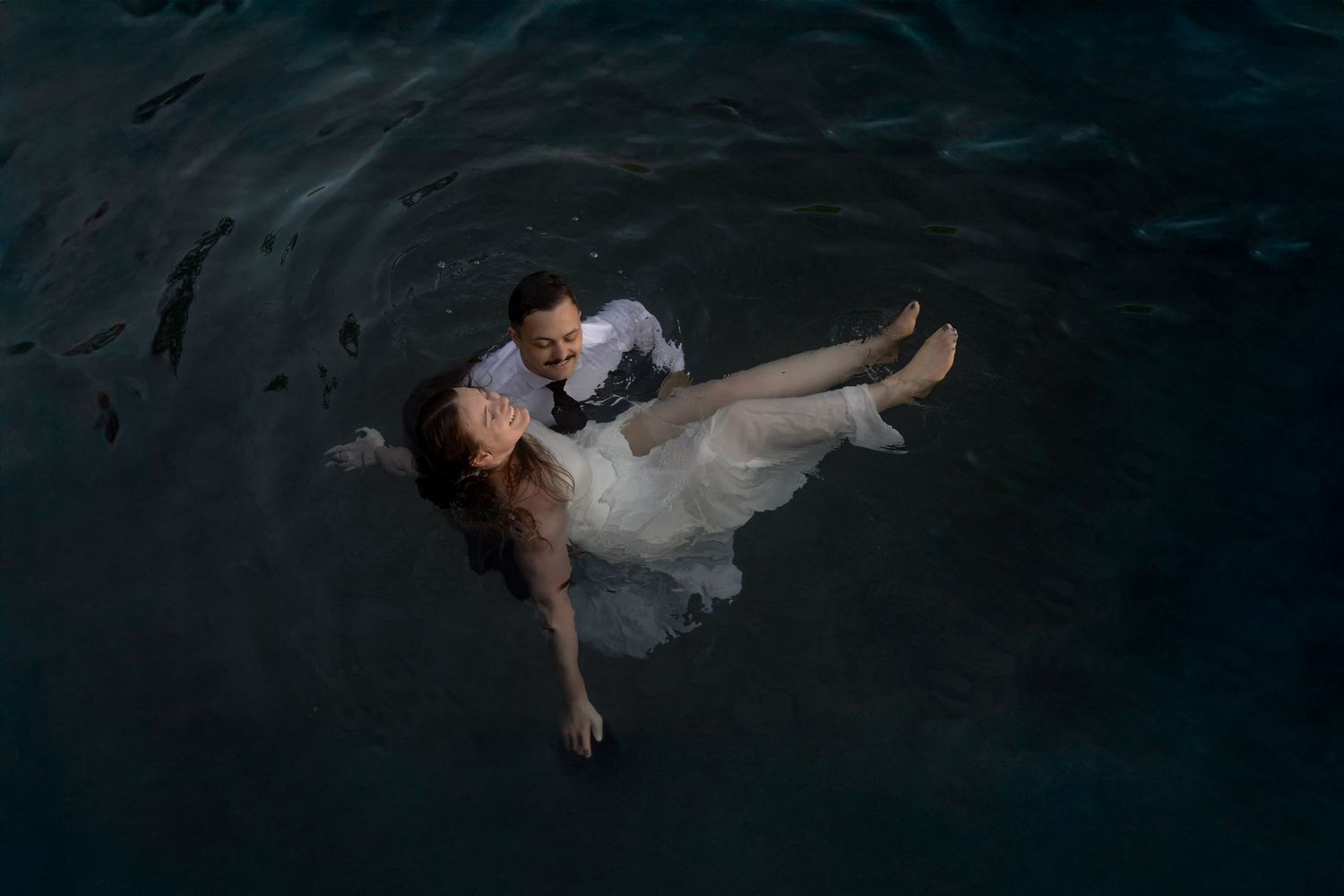 Couple in wedding attire floating together in dark water, bride in white gown, groom in white shirt, aerial view.