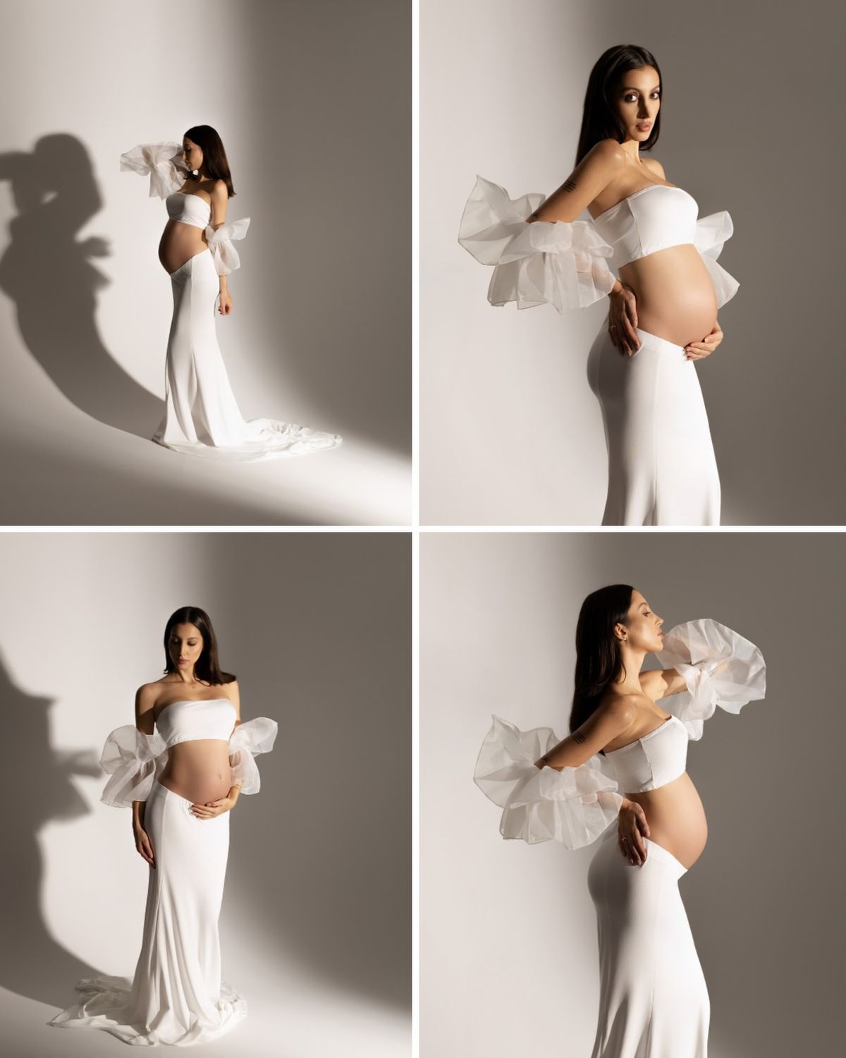 Smiling pregnant woman in white off-shoulder gown with puffy sleeves, cradling baby bump against dark studio background.