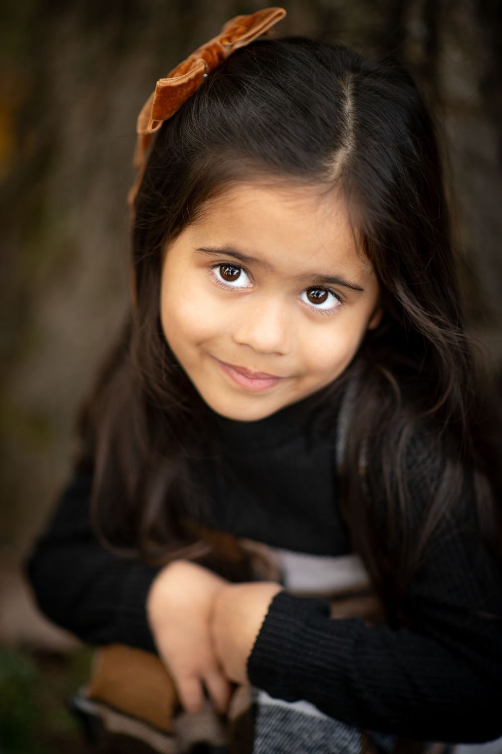 A young girl with long dark hair and a brown bow smiles warmly in an outdoor portrait setting.