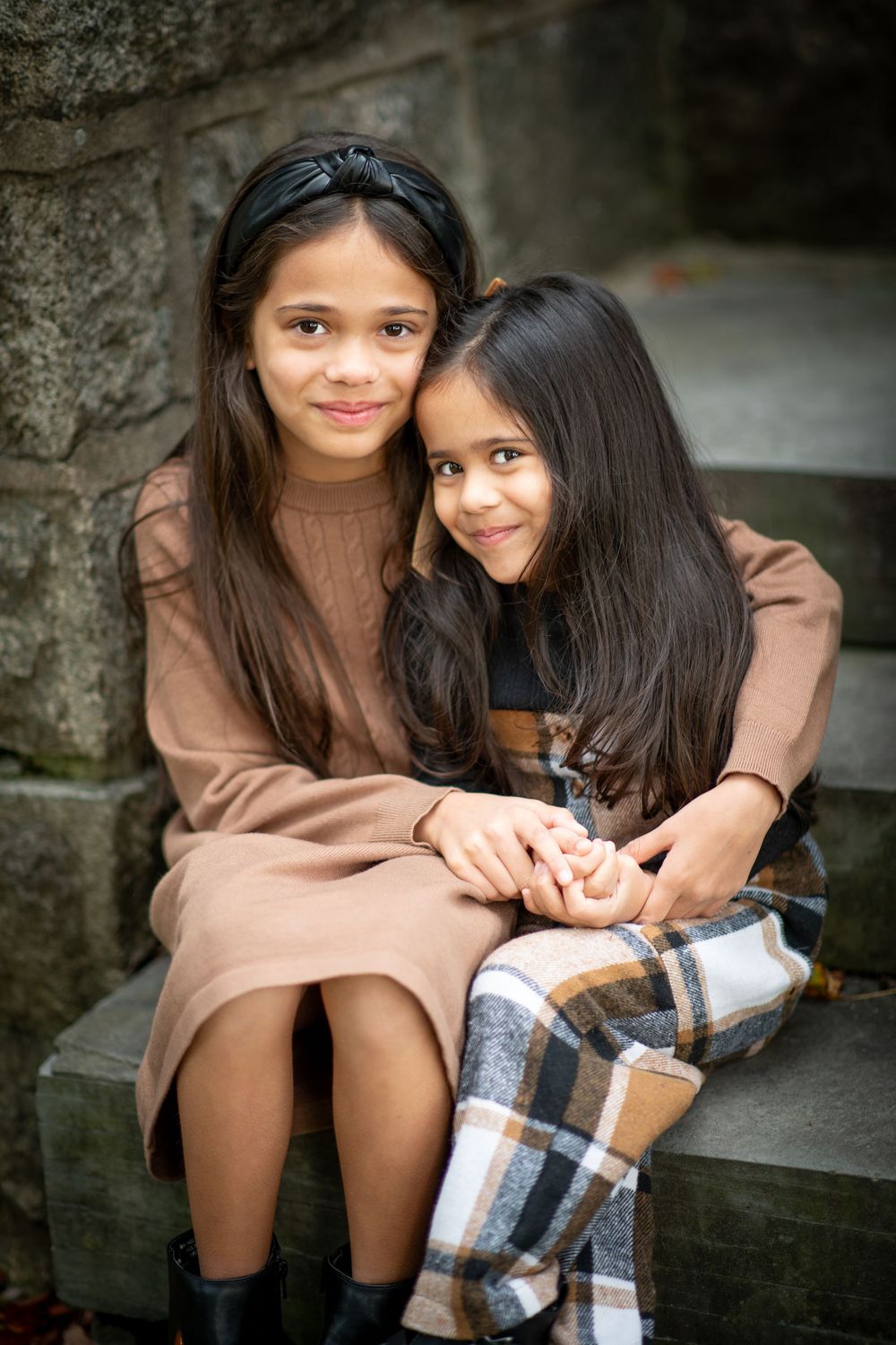 Two young sisters in fall outfits sit together on stone steps, smiling warmly in an outdoor portrait.