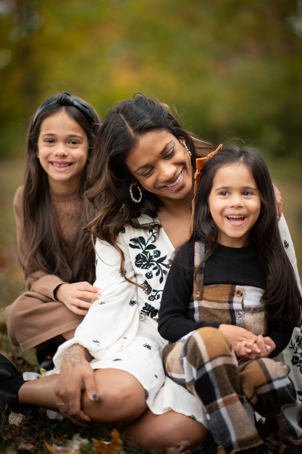 A smiling mother sits outdoors in autumn with her two happy young daughters in a warm fall setting.