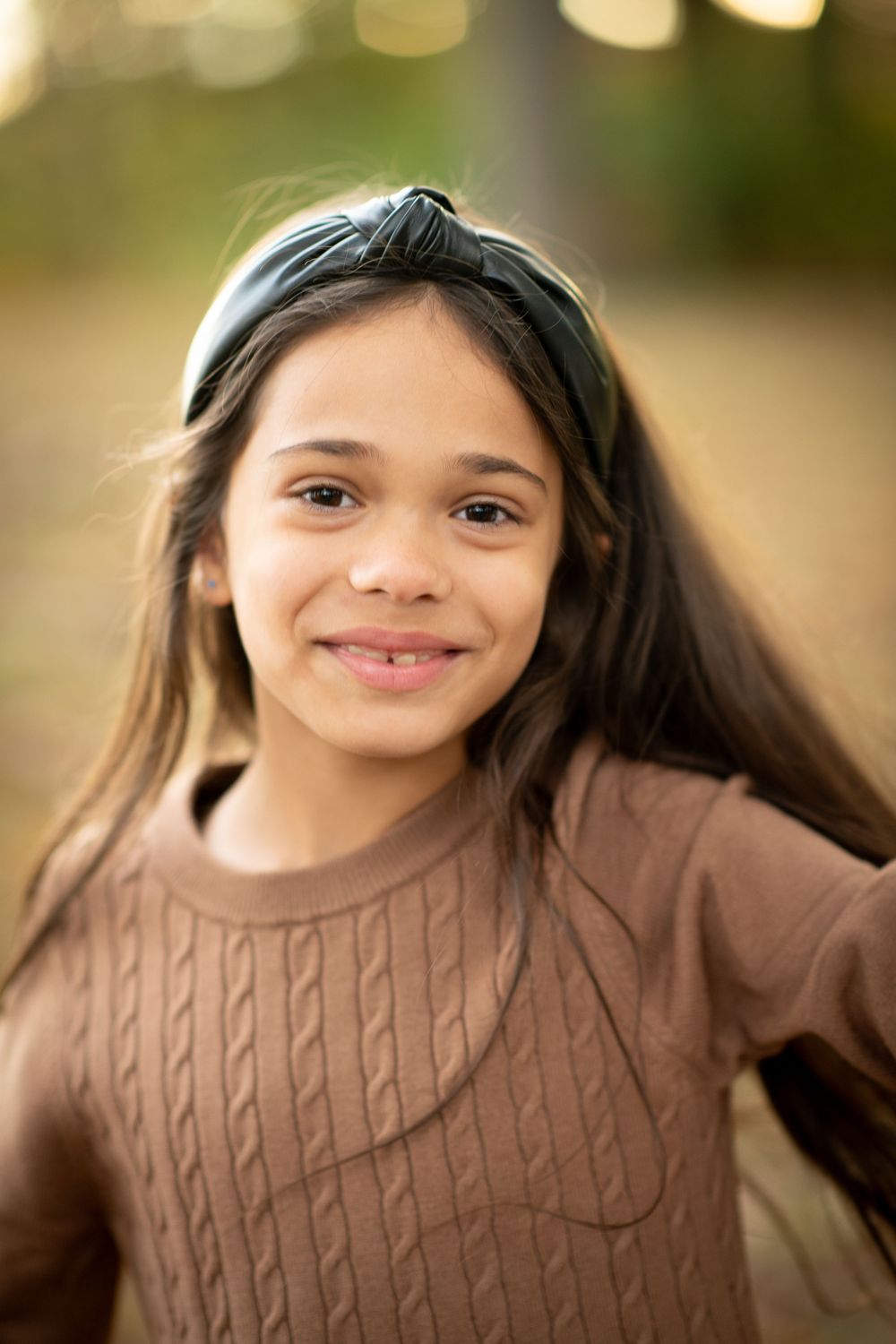 A smiling young girl with long dark hair wearing a green headband and brown cable-knit sweater outdoors.