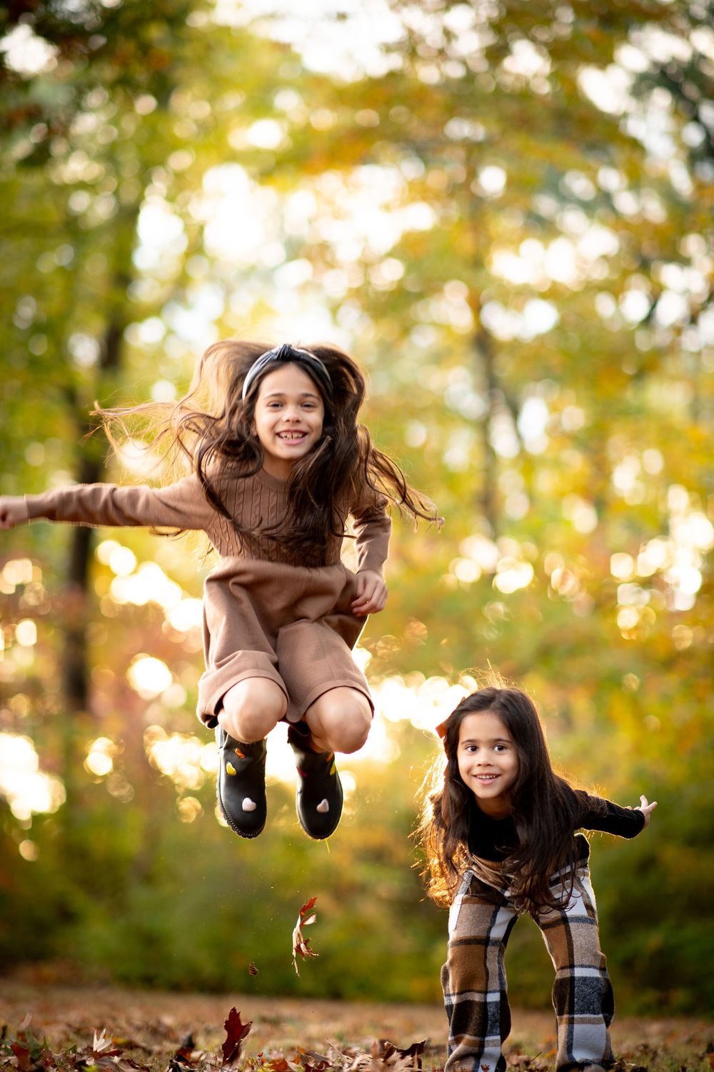 Two young girls playing joyfully in autumn leaves, one leaping in the air, surrounded by golden fall foliage.