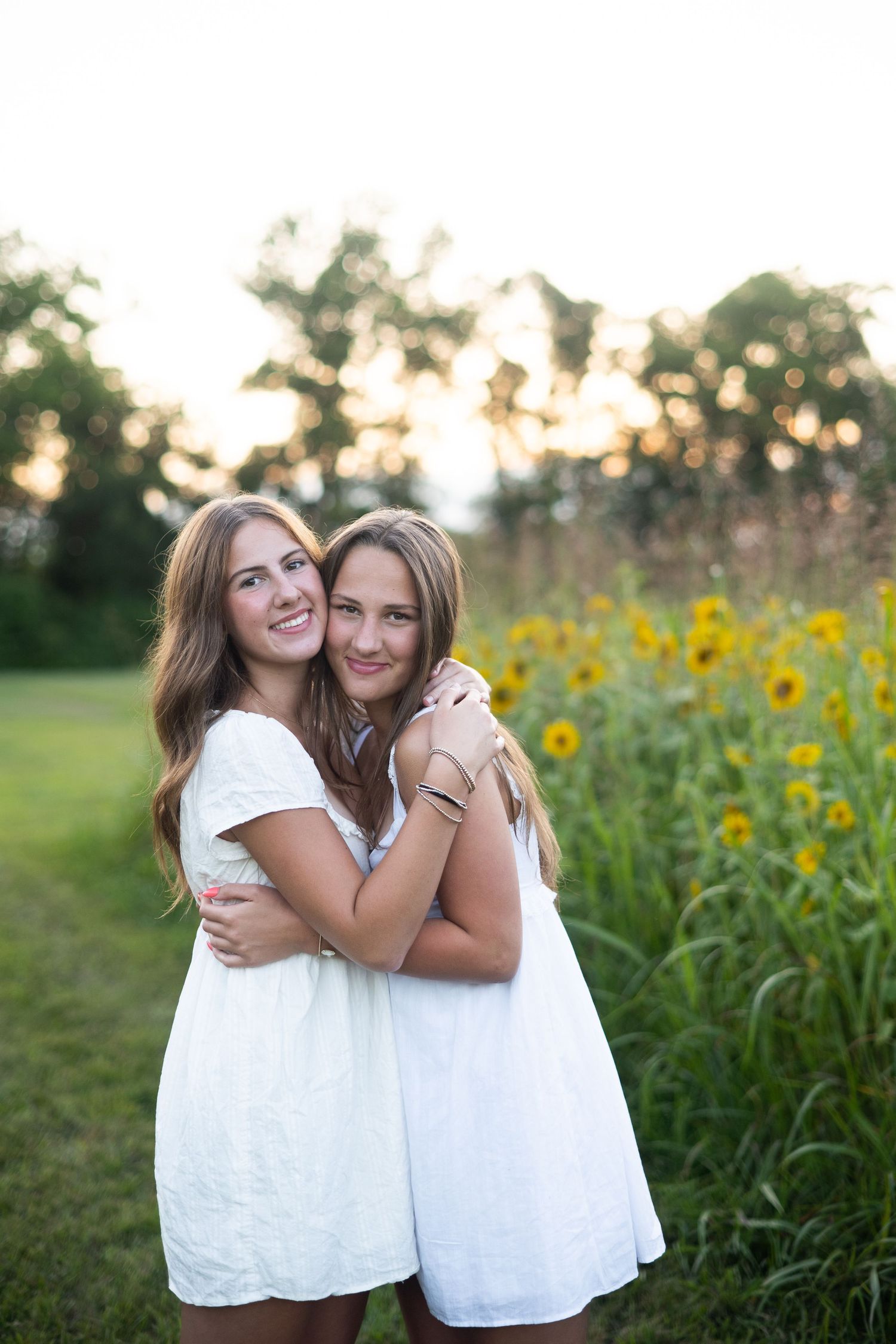 Two young women in white dresses embrace warmly in front of a sunflower field at golden hour.