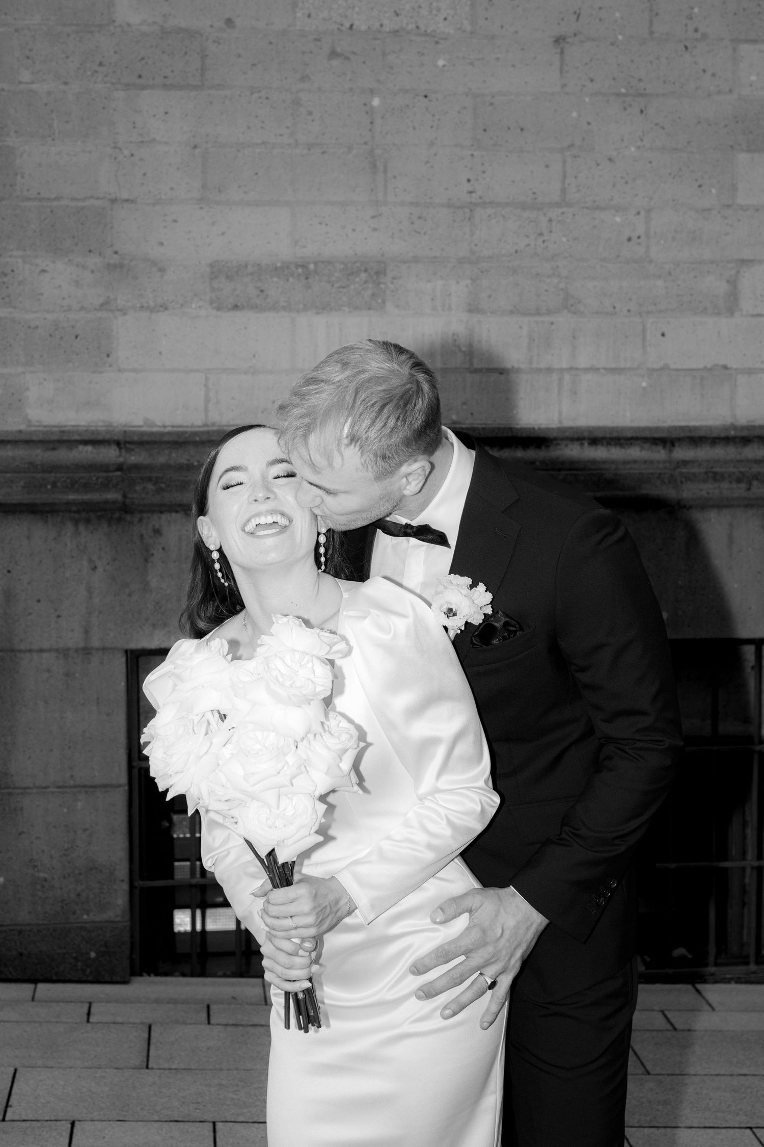 Black and white wedding photo of smiling bride holding bouquet while groom kisses her cheek outdoors.