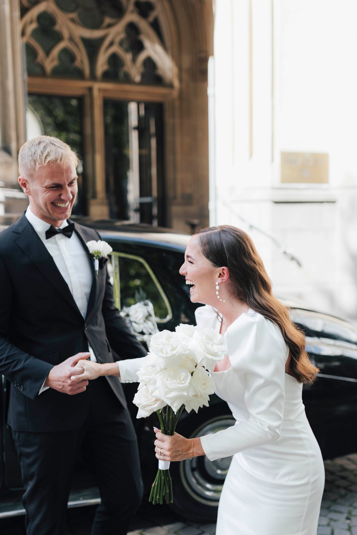 Bride in white dress holds bouquet laughing with groom in black tuxedo outside church near vintage car.