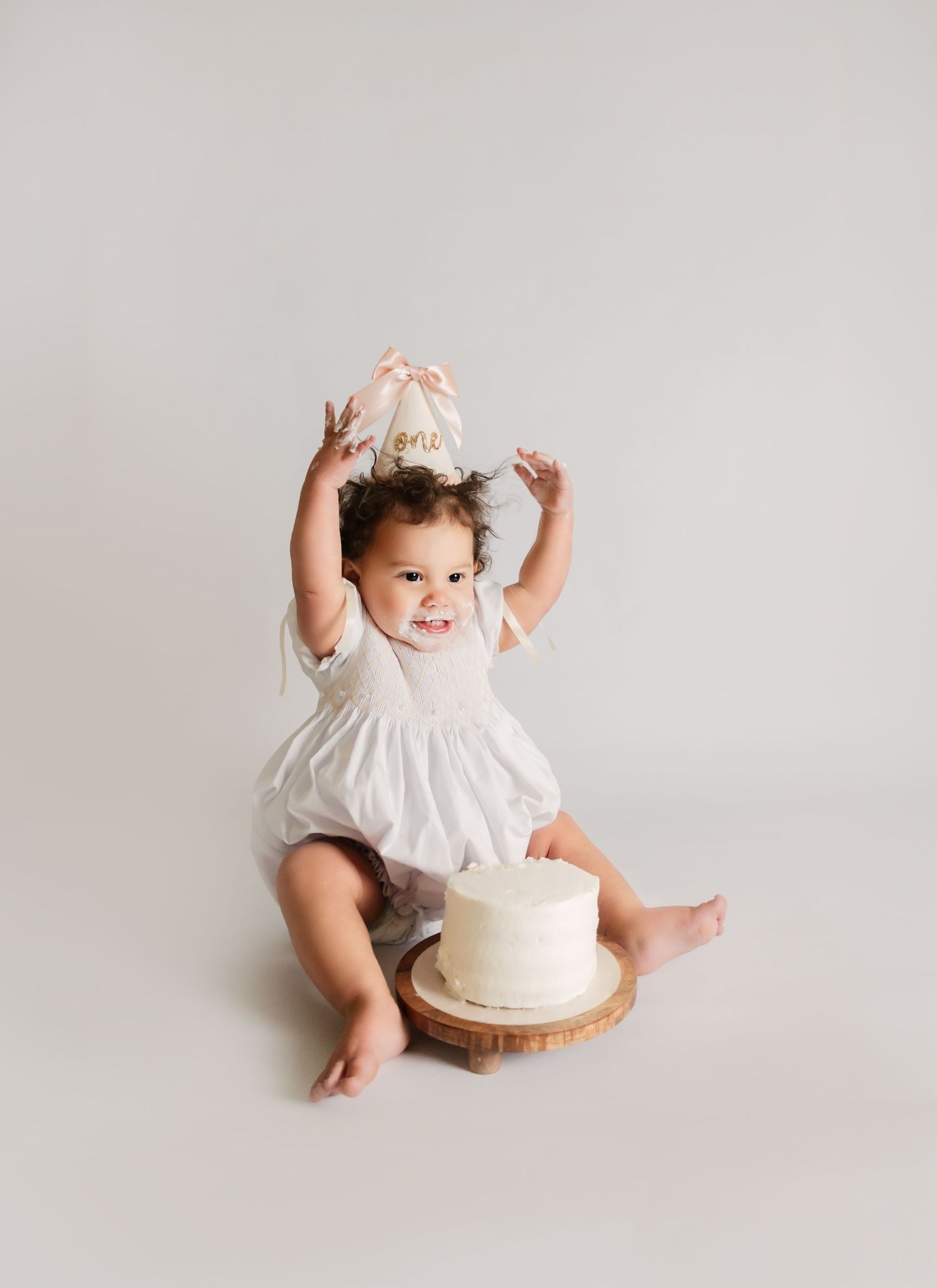Baby girl in white romper wearing a gold ‘one’ party hat celebrates her first birthday with a smash cake.
