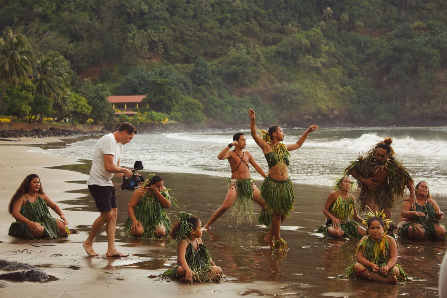 Polynesian dancers in grass skirts perform a traditional hula dance on a tropical beach while a photographer captures the moment.
