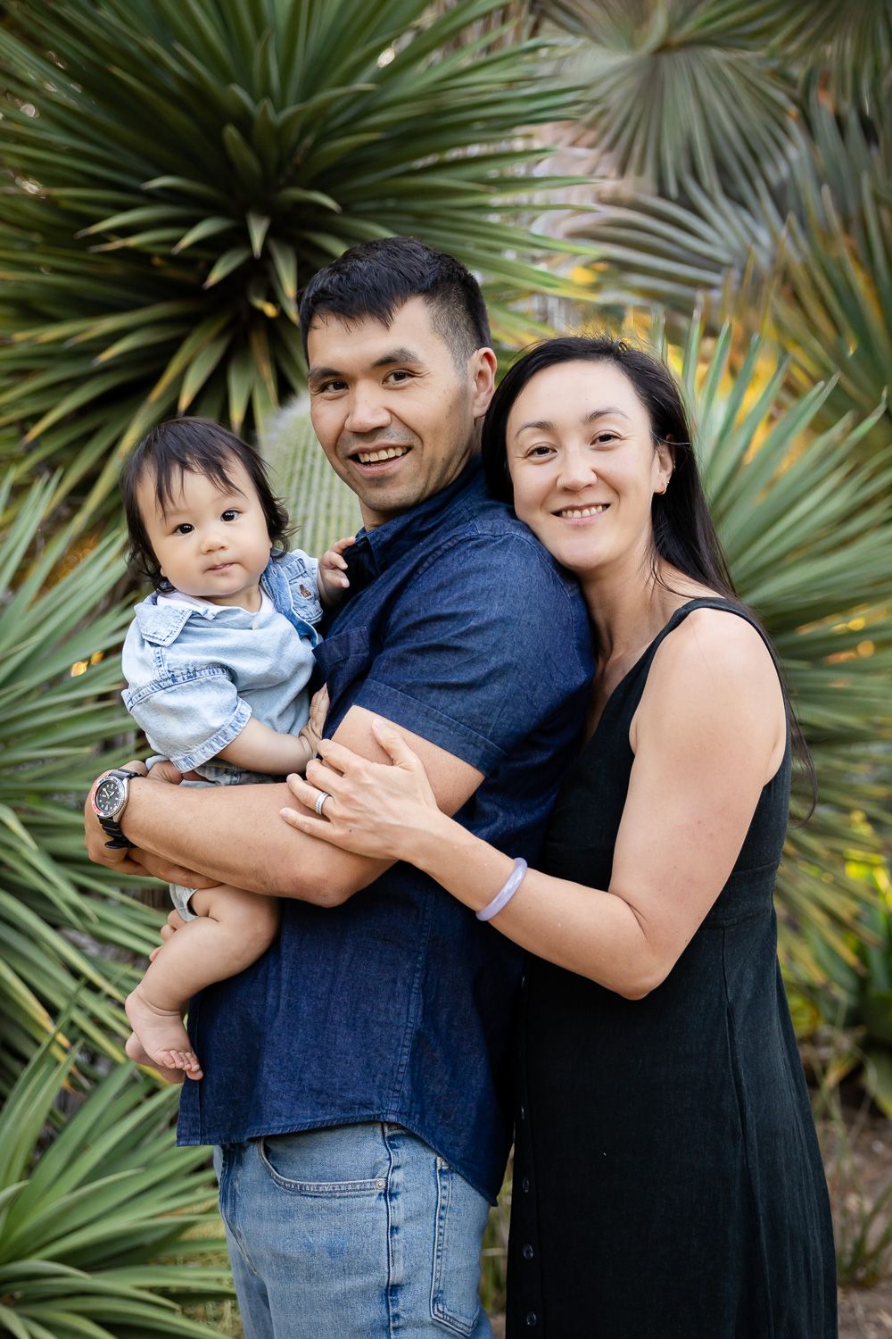 Happy family of three posing outdoors, parents smiling and holding baby in front of palm plants in the Arizona Cactus Garden at Stanford