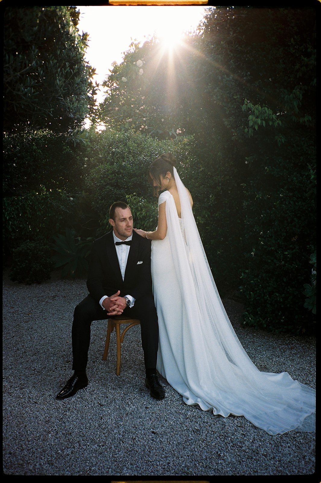 Bride in flowing white gown and veil stands beside groom in black tuxedo seated on chair, bathed in golden sunlight.
